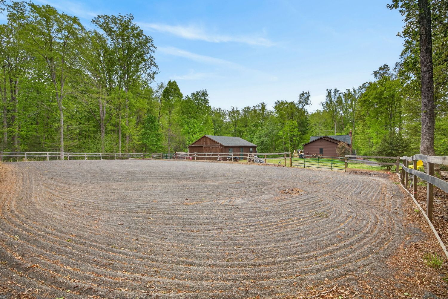 Expansive gravel equestrian riding arena surrounded by lush trees with rustic barn buildings in the background.