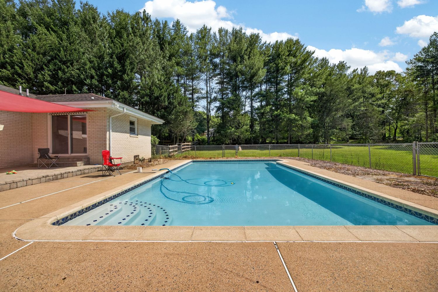 Rectangular backyard swimming pool with clear blue water, surrounded by a concrete deck, small pool house, and lush green trees.