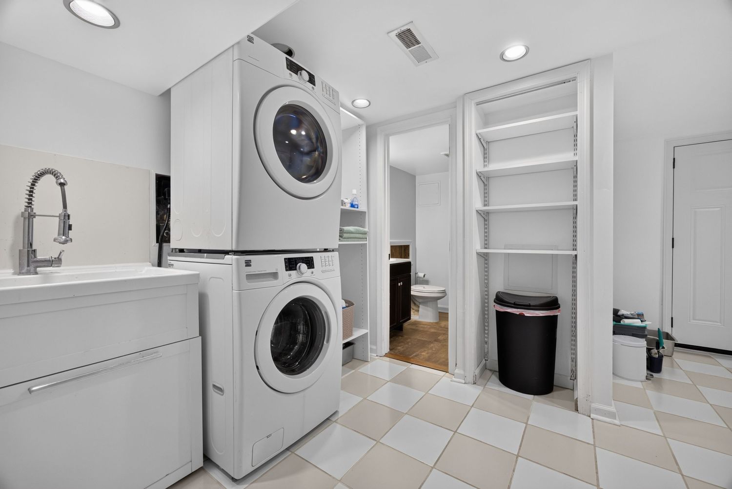 Laundry room with stacked washer and dryer, utility sink, checkered tile floor, and open shelving near a bathroom.