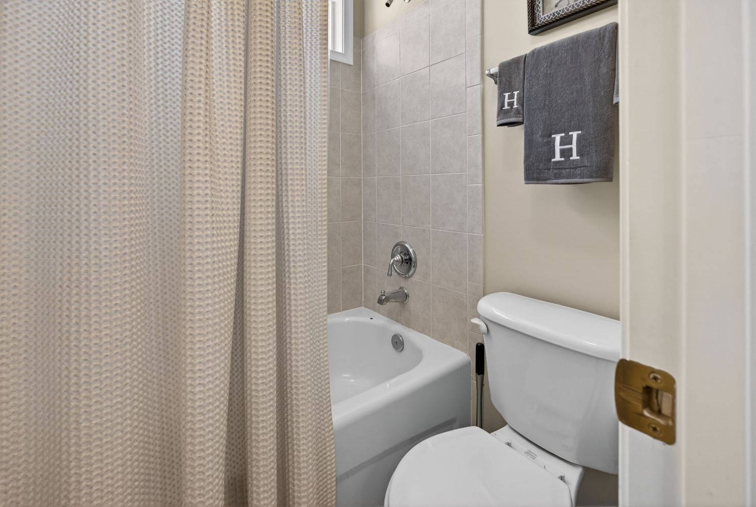 Compact bathroom featuring a bathtub with beige curtain, white toilet, and neutral tiled walls with monogram towel decor.
