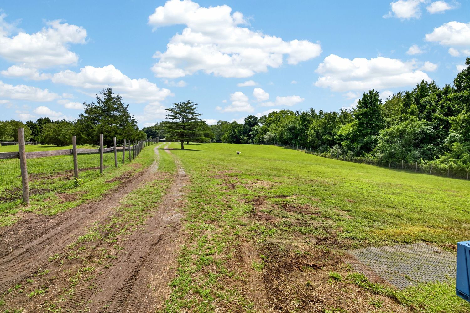 Rural farm property featuring a dirt path along wooden fence posts, lush green pasture, and tree line under a partly cloudy sky.