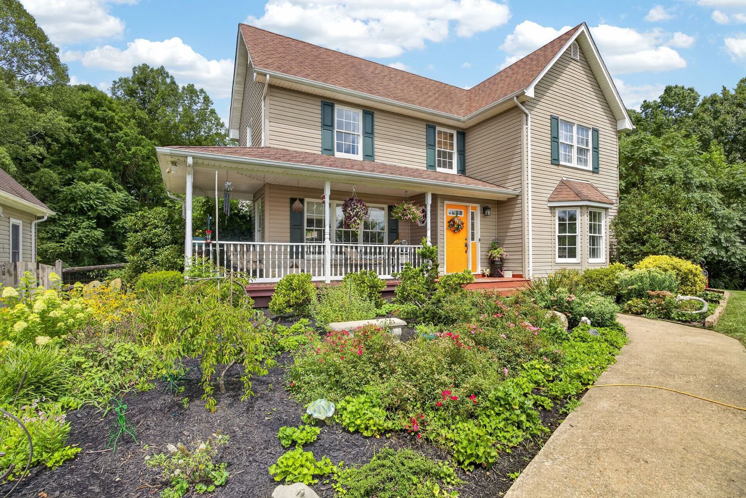 Charming two-story home with wrap-around porch, orange front door, and lush landscaped garden in a wooded setting.