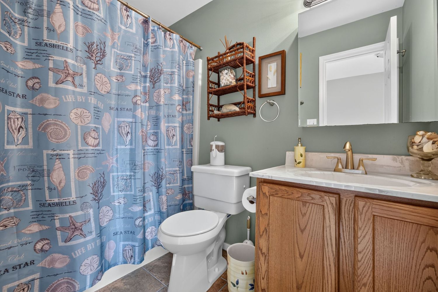 Coastal-themed bathroom with blue seashell shower curtain, wood vanity with gold fixtures, white toilet, and decorative shelving.