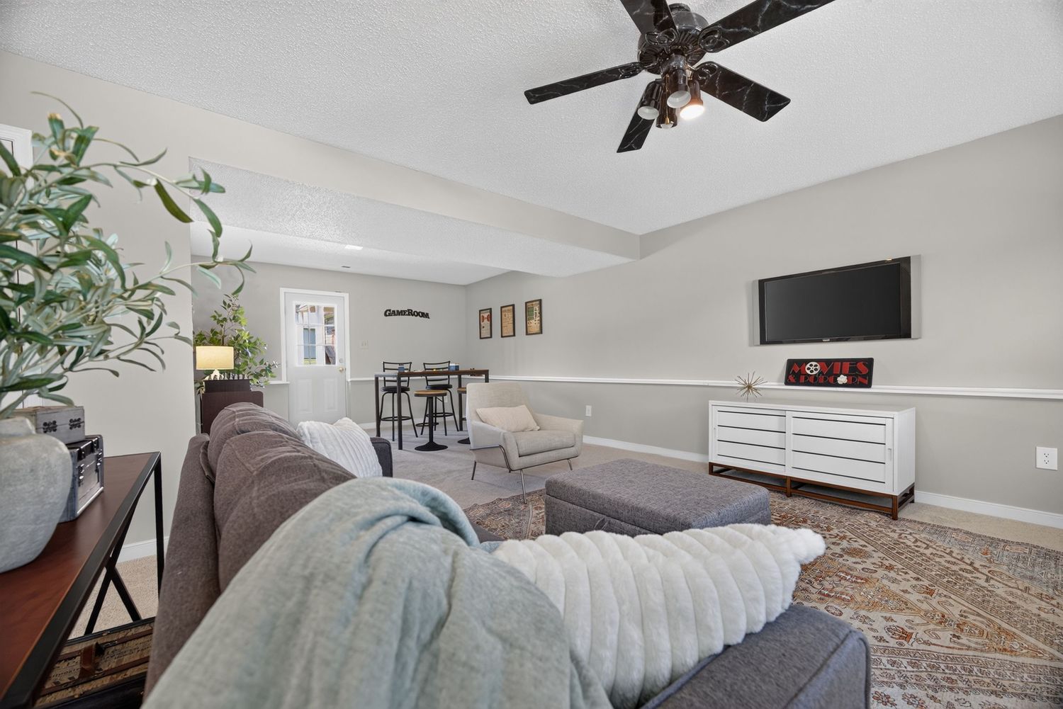 Cozy living room with gray sectional sofa, ceiling fan, wall-mounted TV, white dresser, and warm hardwood floors.
