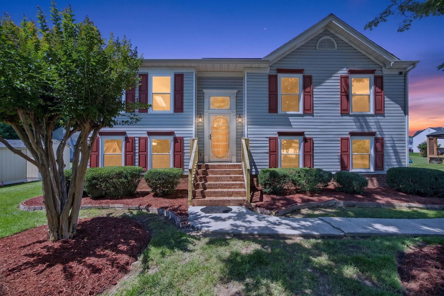 Two-story gray colonial home with red shutters, brick steps, and lush landscaping photographed at dusk.