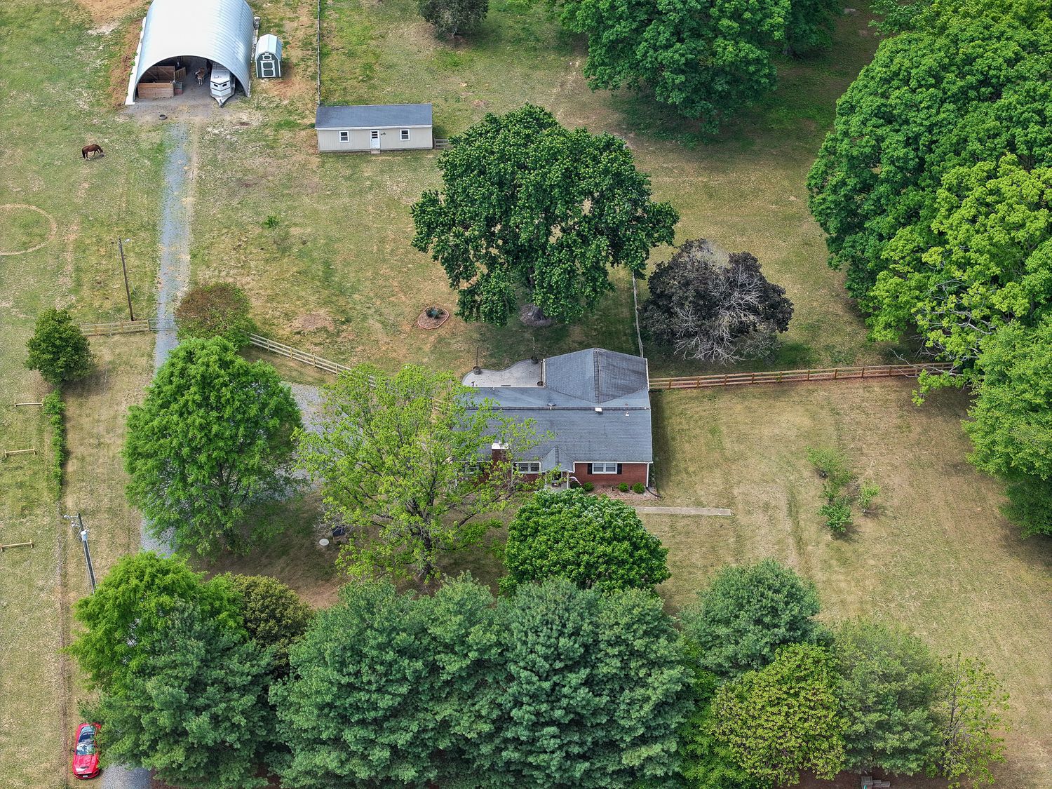 Aerial view of a rural property featuring a small house, metal barn, and lush green trees on a spacious grassy lot.