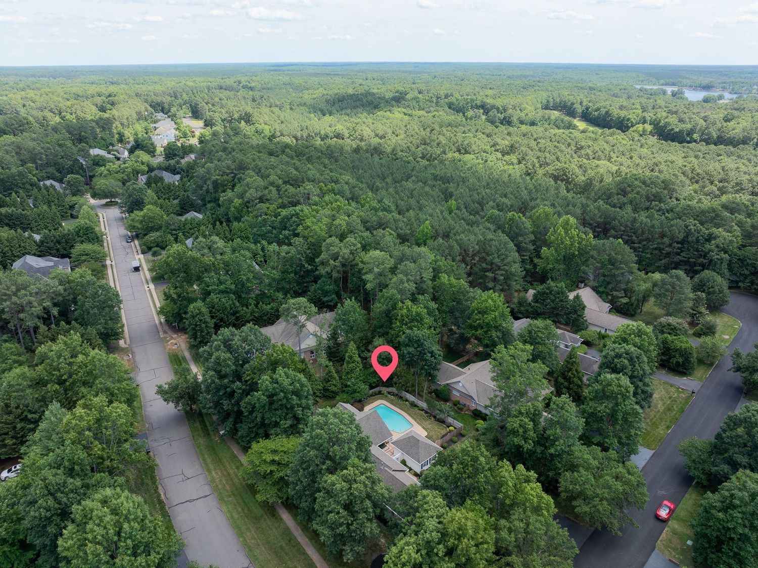 Aerial drone view of a suburban home with a pool surrounded by lush green trees and nearby roads in a wooded neighborhood.