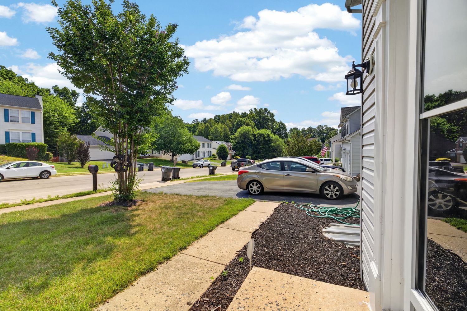 Suburban neighborhood street view with parked cars, green lawn, and mature trees on a sunny summer day.