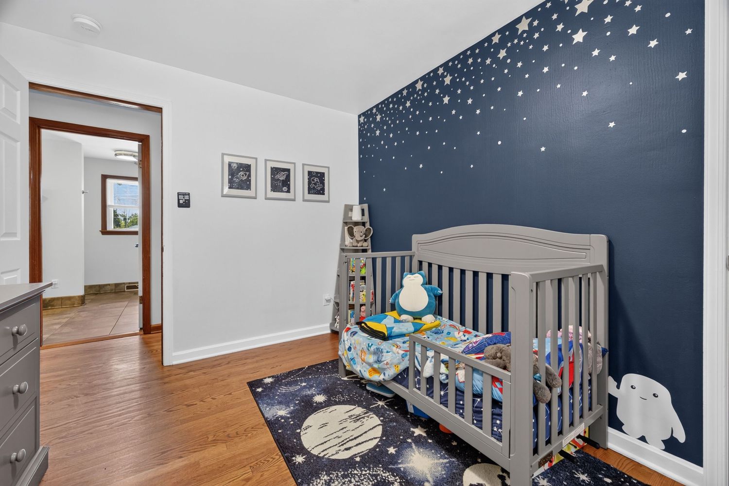 Cozy nursery with navy star-wall accent, gray crib, space-themed rug, and hardwood floors.