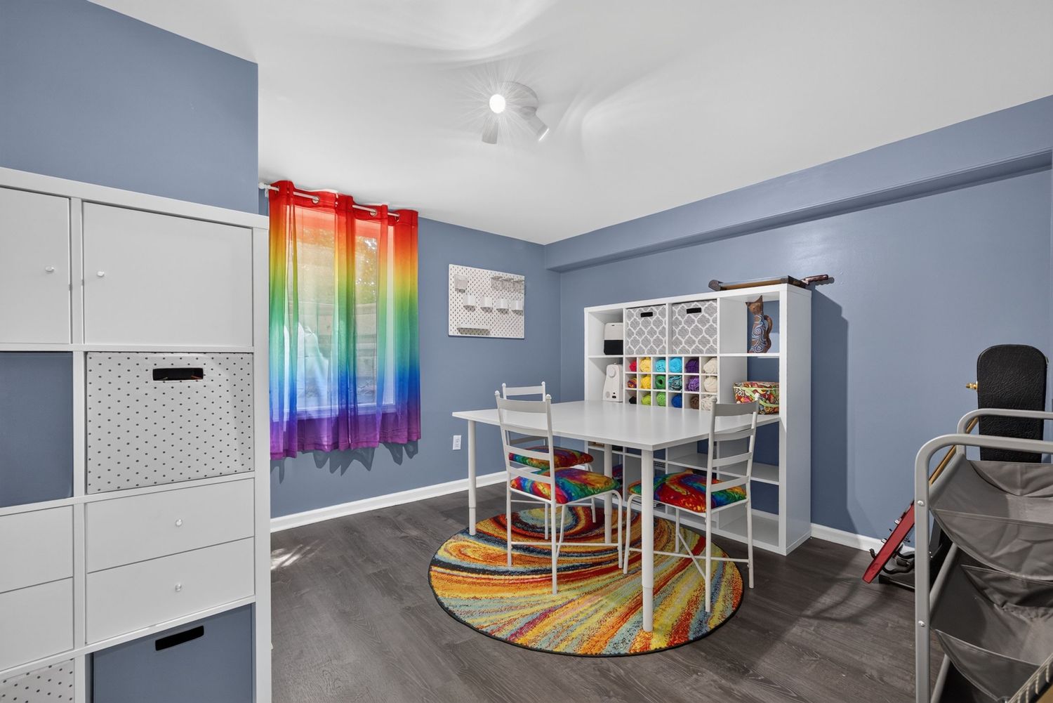 Bright craft room with blue walls, rainbow curtains, white storage cubes, colorful rug, and a table with chairs on dark hardwood floors.
