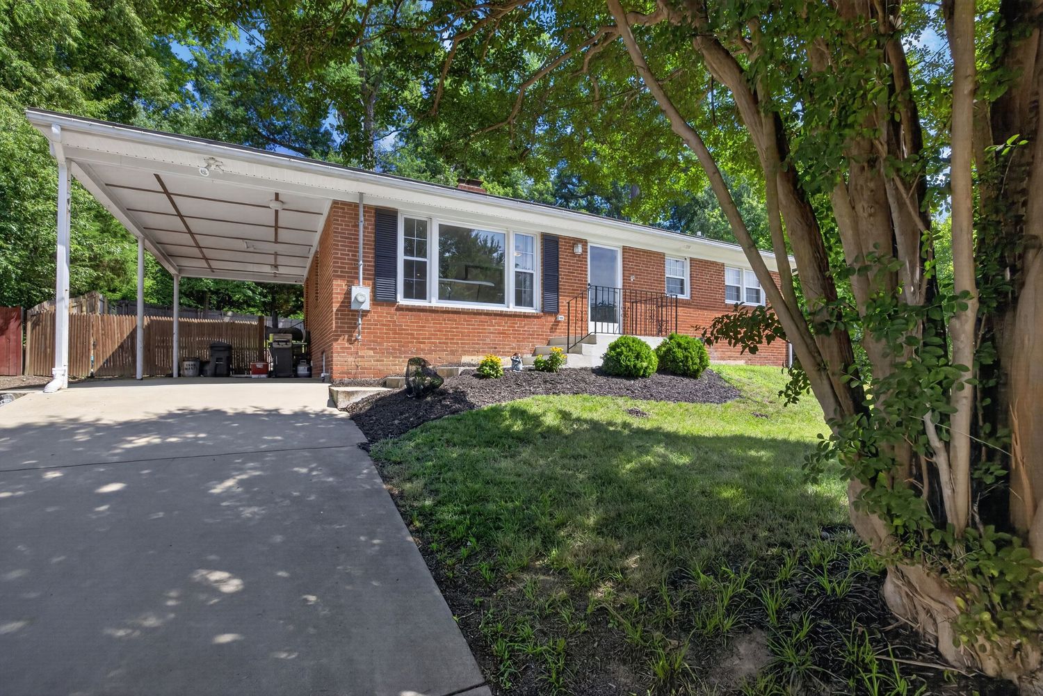 Brick ranch home with carport, lush green trees, manicured lawn, and concrete driveway on a sunny day.