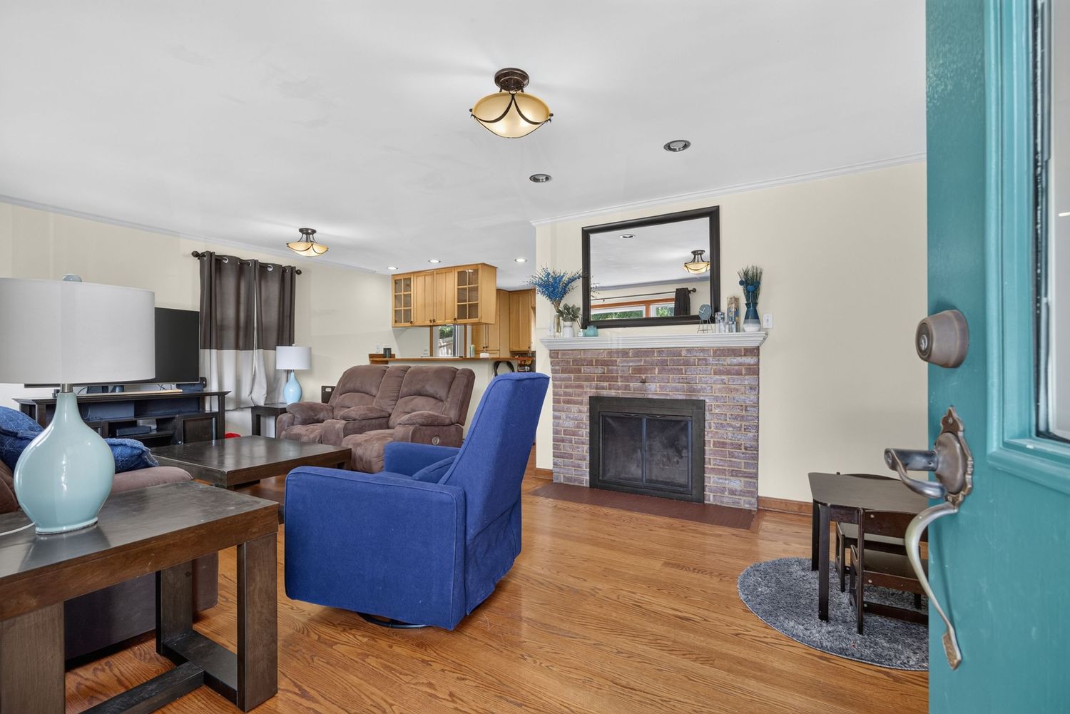 Bright living room with hardwood floors, blue armchair, brick fireplace, and open view to kitchen with wood cabinetry.