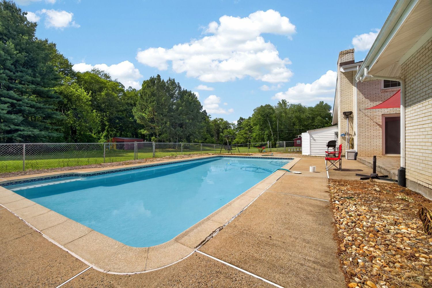 Rectangular backyard swimming pool with blue water surrounded by concrete patio, lush green trees, and adjacent two-story home.
