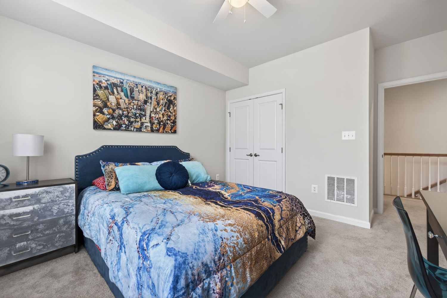 Cozy bedroom with blue upholstered bed, colorful cityscape artwork, ceiling fan, and neutral carpet flooring.