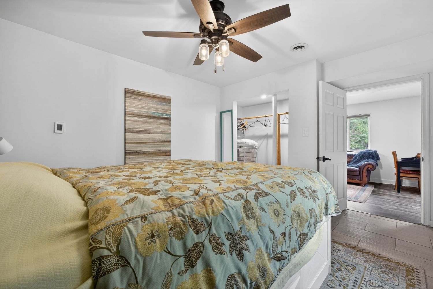 Bright bedroom with floral bedspread, ceiling fan, hardwood accent wall, and open doorway leading to ensuite and sitting room.