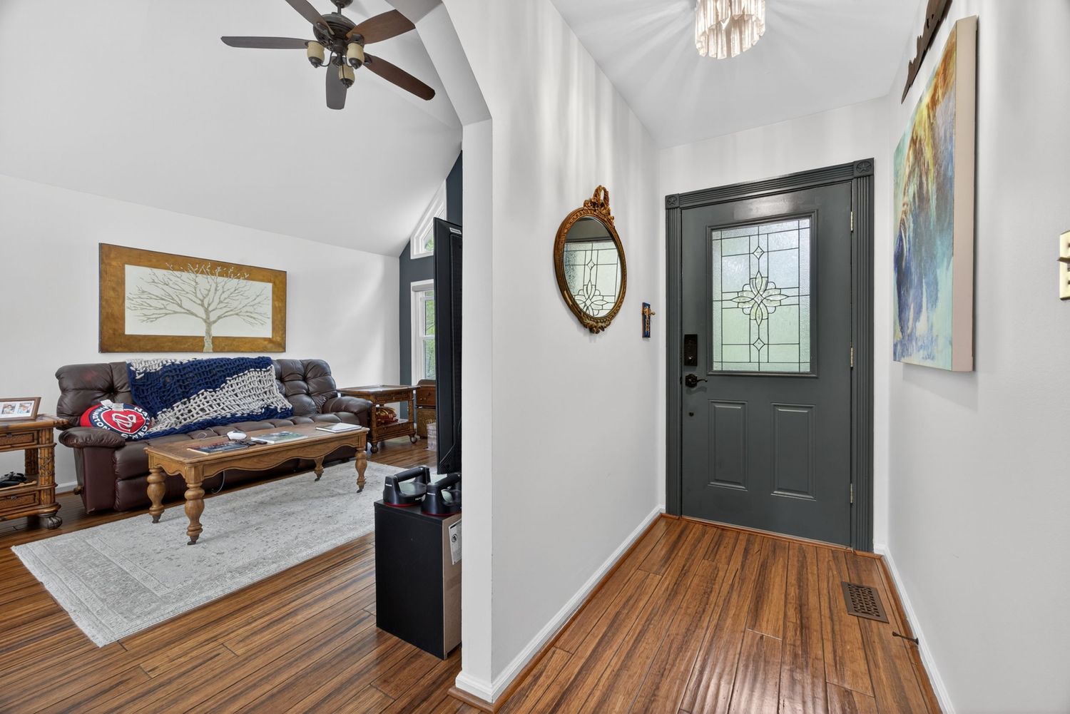 Bright entryway with dark front door, stained glass, hardwood floors, and open view into cozy living room with ceiling fan.