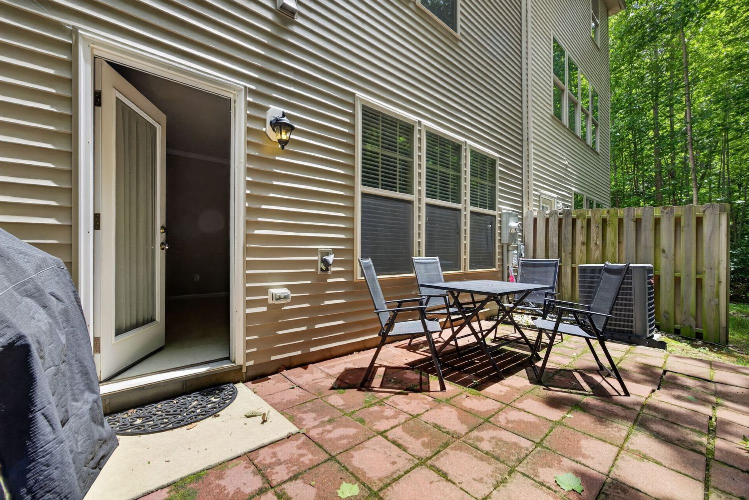 Cozy patio with brick pavers, outdoor dining set, and lush wooded backdrop beside a beige townhome.