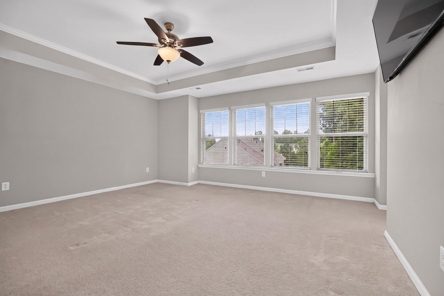 Spacious bedroom with tray ceiling, ceiling fan, large windows, and neutral carpet flooring in a modern home.