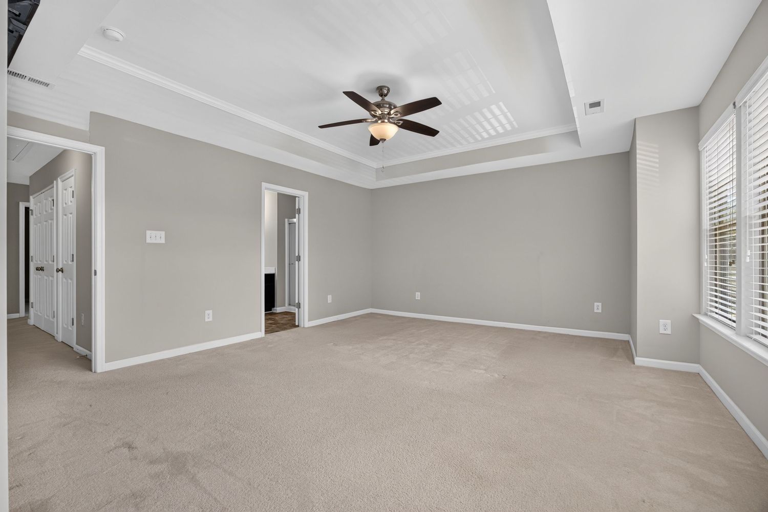 Spacious bedroom with tray ceiling, ceiling fan, neutral carpet, gray walls, and natural light from large windows.