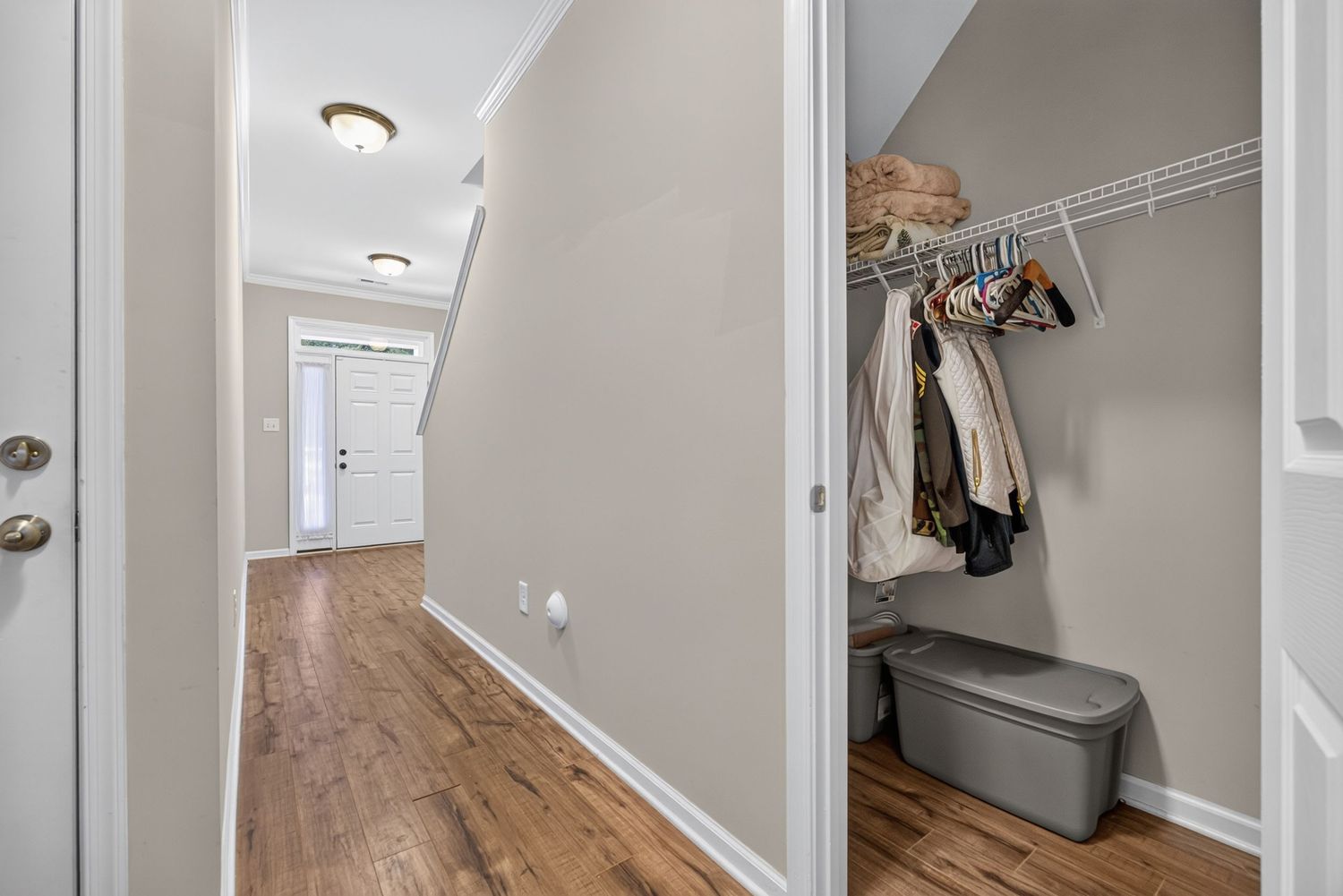 Hallway with hardwood floors leading to front door, adjacent open closet with hanging clothes and storage bench.