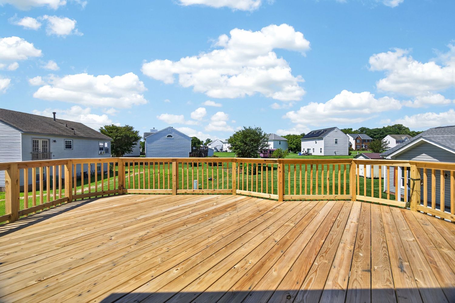 Spacious wooden deck with railings overlooking a residential neighborhood under a bright blue sky with fluffy white clouds.