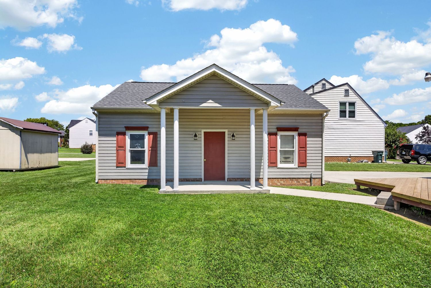 Gray cottage-style home with red shutters, covered front porch, and lush green lawn on a sunny day.