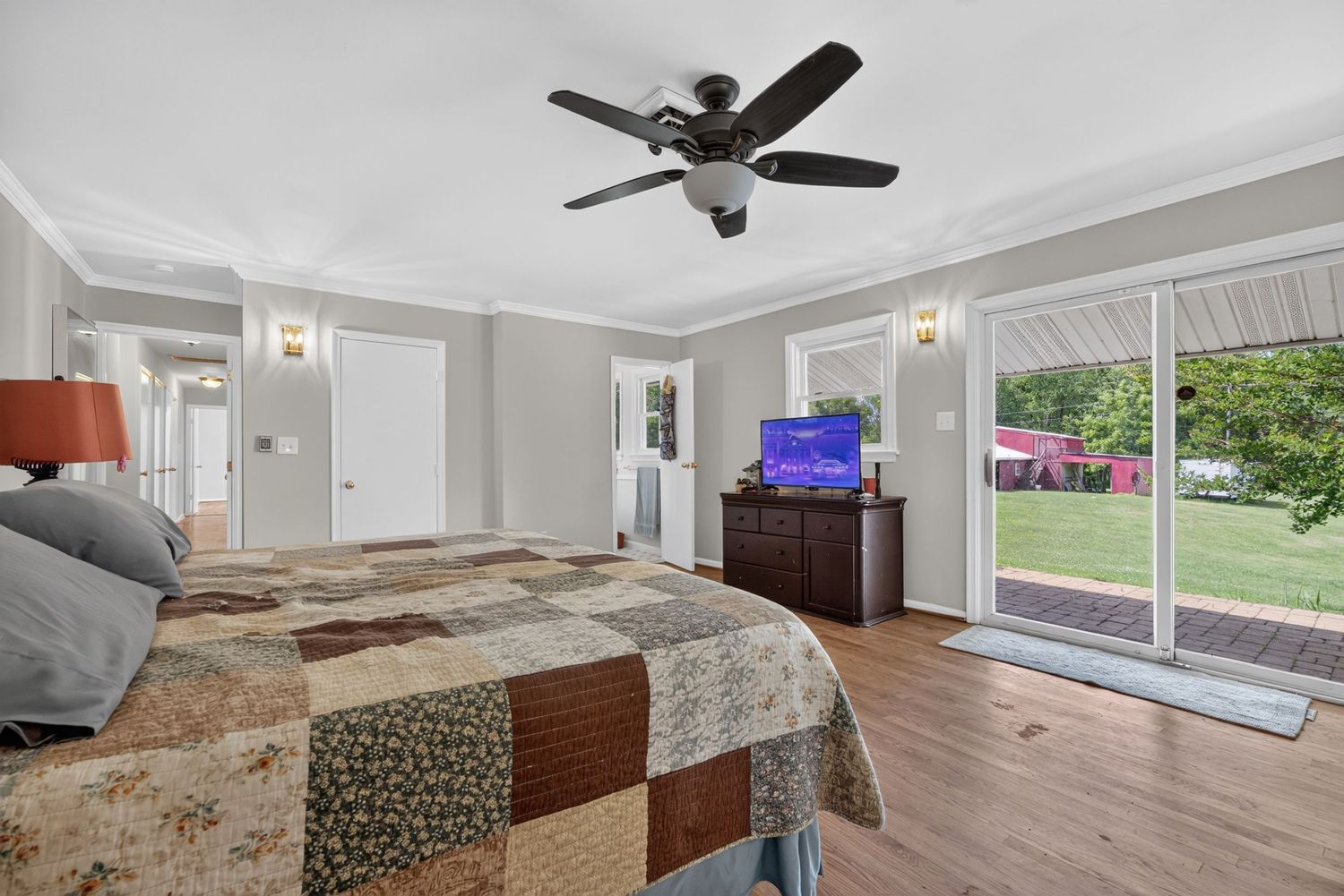 Bright bedroom with hardwood floors, ceiling fan, patchwork quilt bed, dresser with TV, and sliding glass doors to a green backyard.