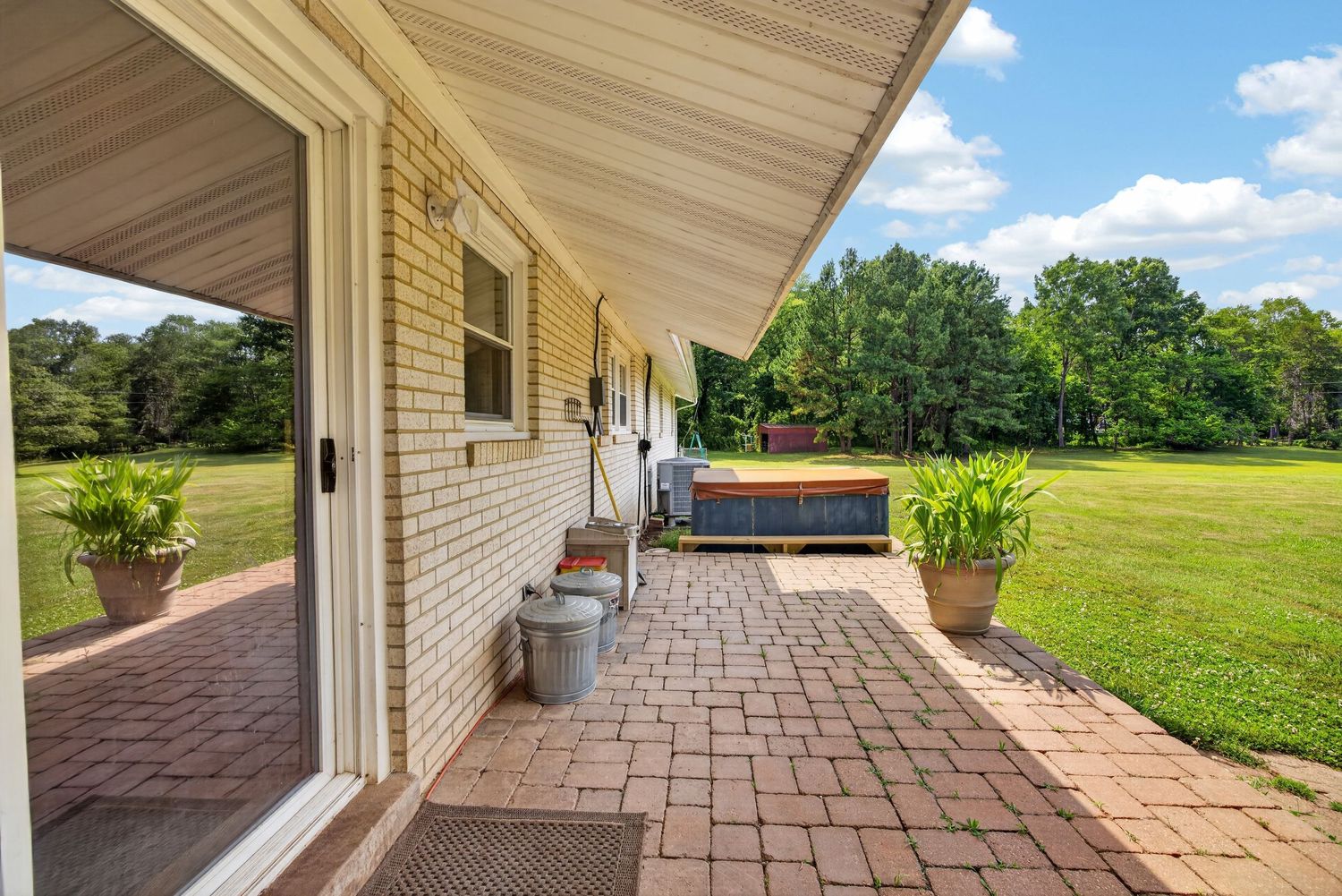Brick home patio with hot tub, potted plants, and paver walkway overlooking lush green lawn and trees.