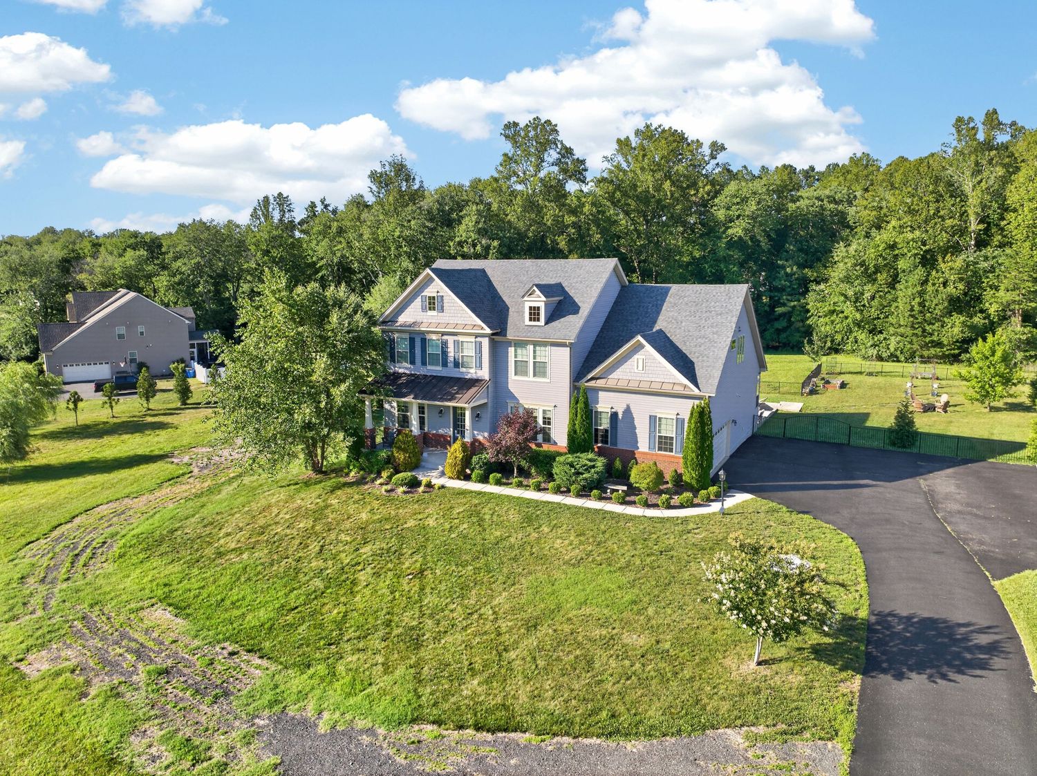 Aerial view of a large two-story colonial home with gray roof, wrap-around porch, and lush green lawn surrounded by trees.