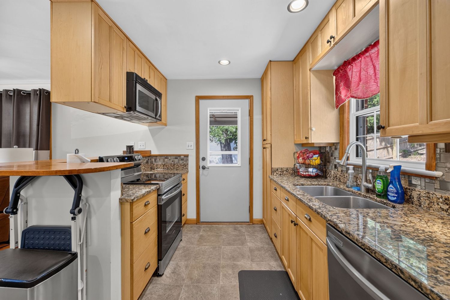 Galley kitchen with light wood cabinets, granite countertops, stainless steel appliances, and a window above the sink.