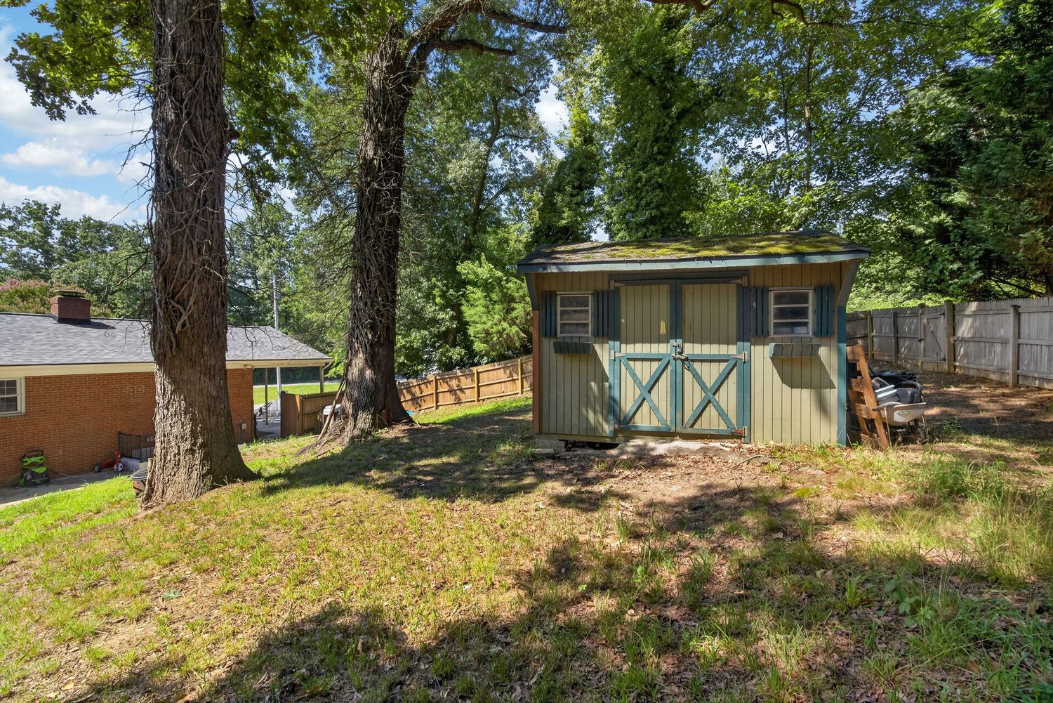 A charming wooden shed with blue trim sits in a lush green backyard shaded by large oak trees.