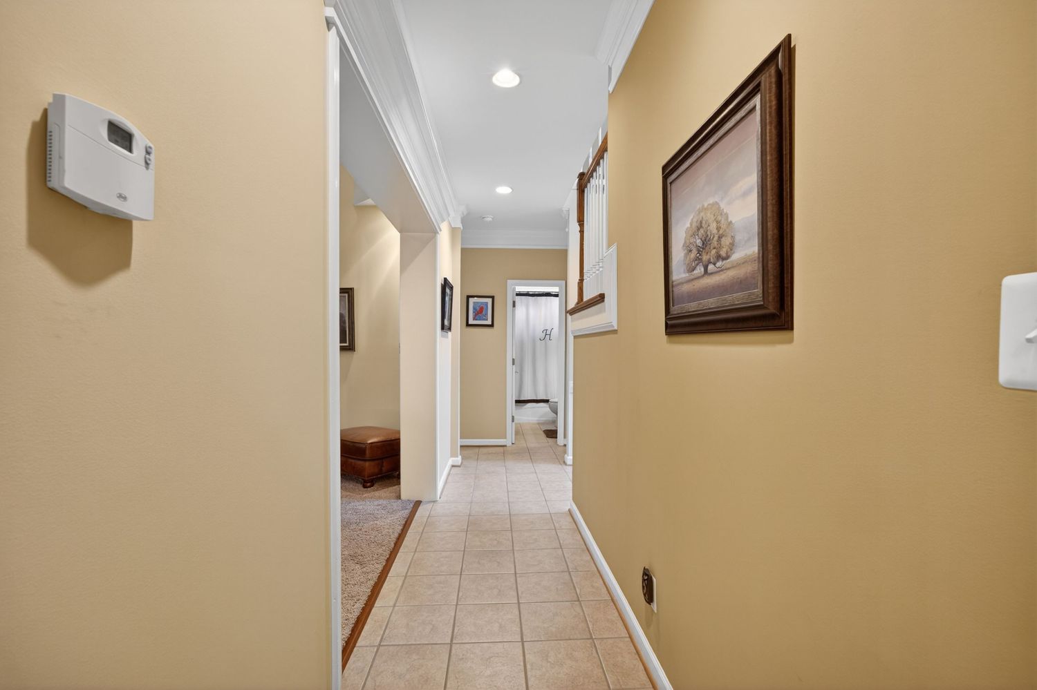 Bright hallway with beige walls, tile flooring, crown molding, framed art, and recessed lighting leading to rooms beyond.