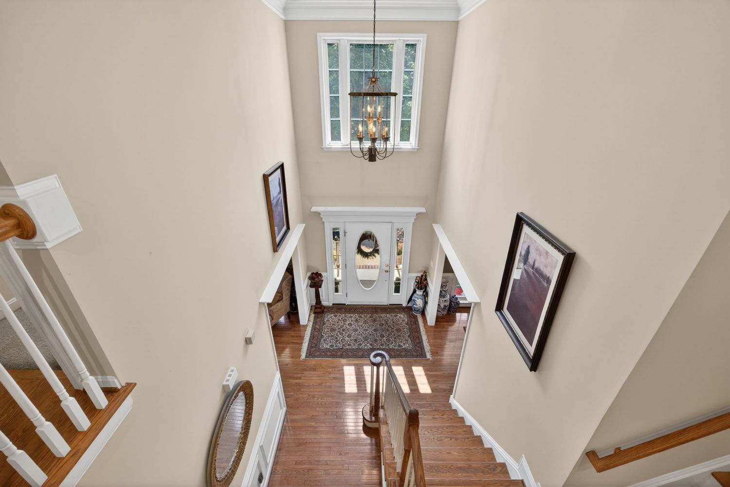 Elegant two-story foyer with chandelier, white staircase, hardwood floors, and framed artwork viewed from above.