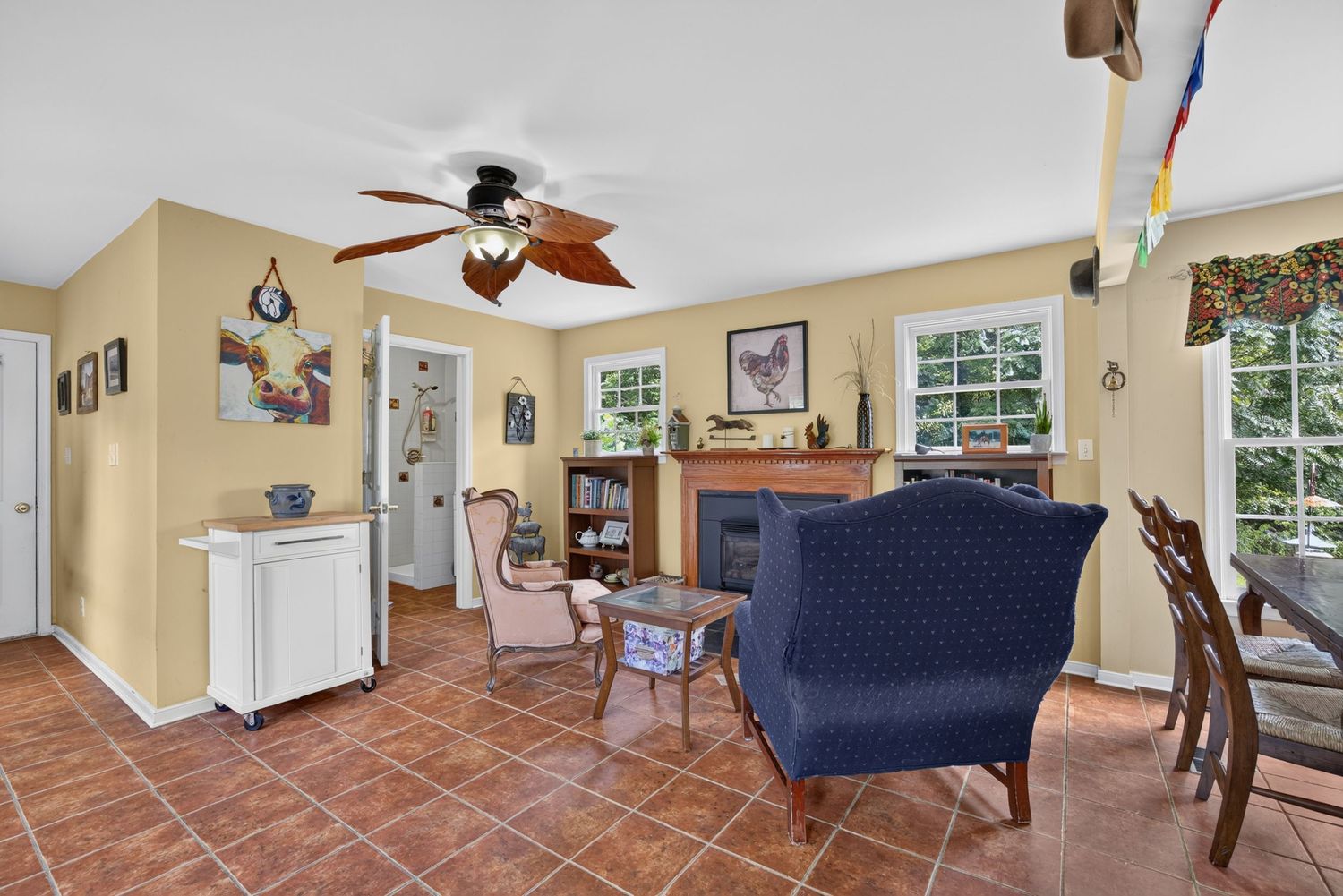 Cozy living room with blue armchair, ceiling fan, fireplace, and large windows in a warm yellow-painted home.