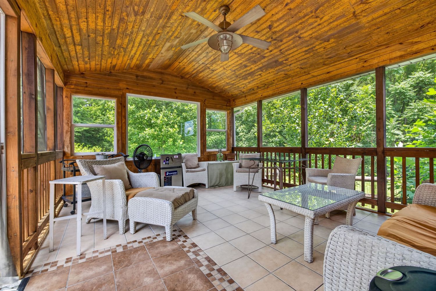 Screened porch with wood ceiling, ceiling fan, white wicker furniture, tile floors, and lush green tree views.