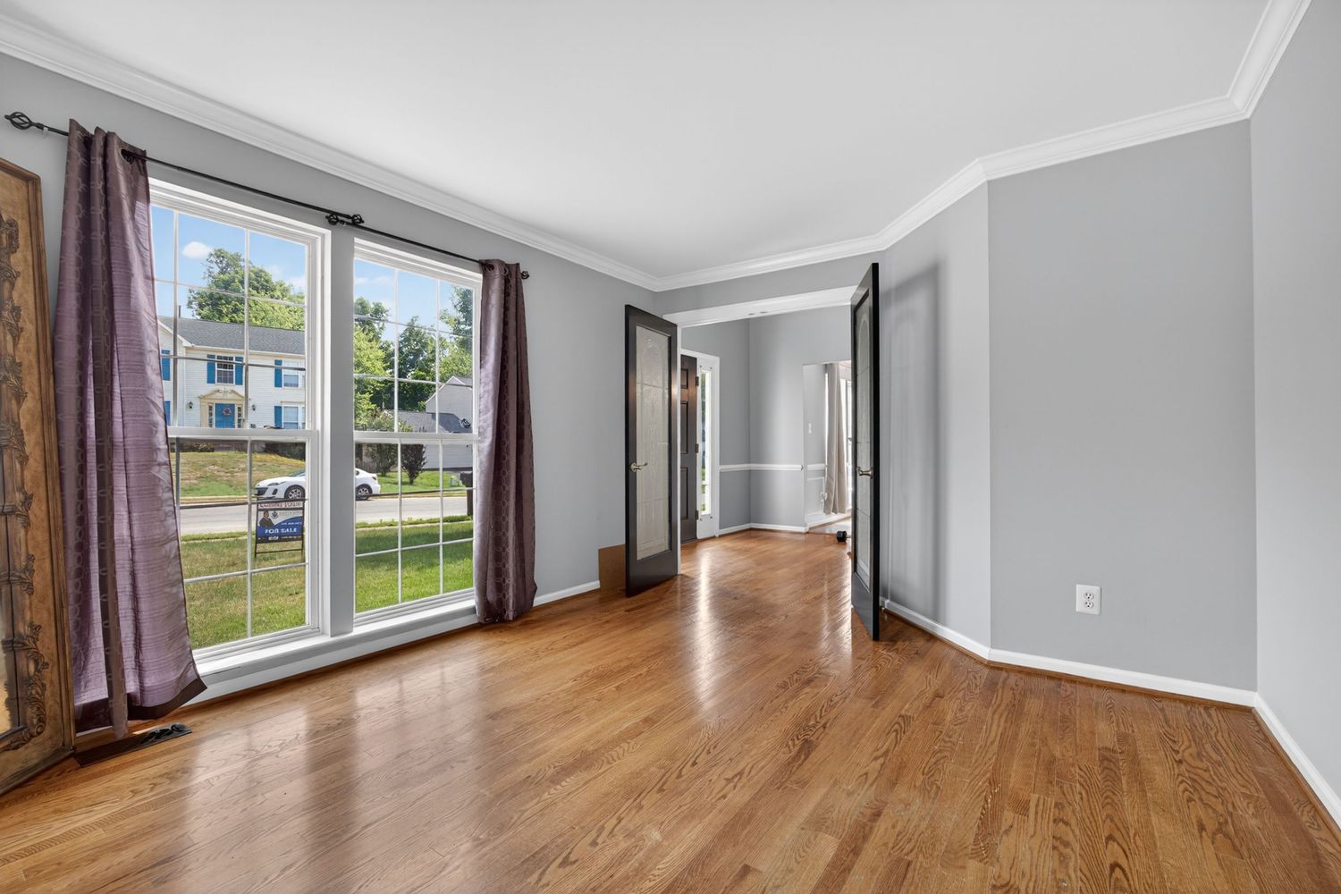Bright empty living room with hardwood floors, large windows, crown molding, and neutral gray walls.