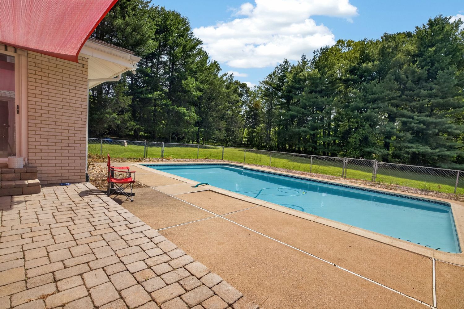 Outdoor rectangular swimming pool with brick patio, red chairs, and lush green trees in background on sunny day.