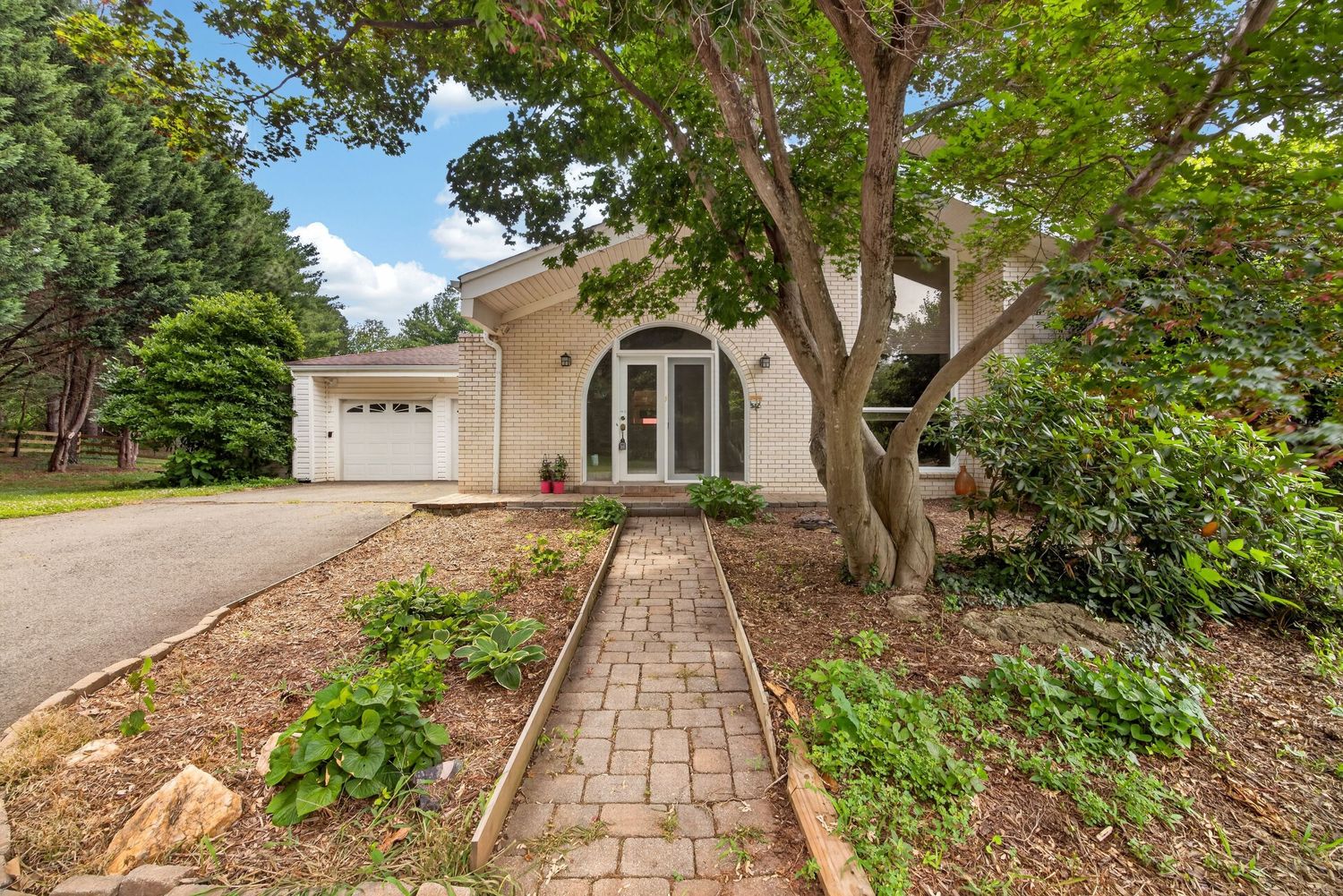 Charming home exterior with brick pathway leading to arched entry, white garage, and lush mature trees surrounding the property.