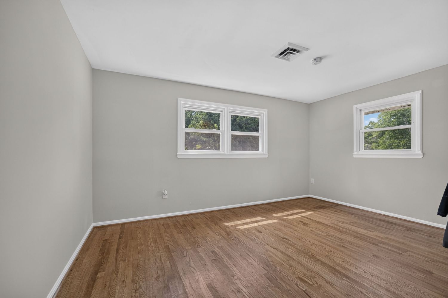 Empty room with light gray walls, hardwood floors, and two windows allowing natural light.