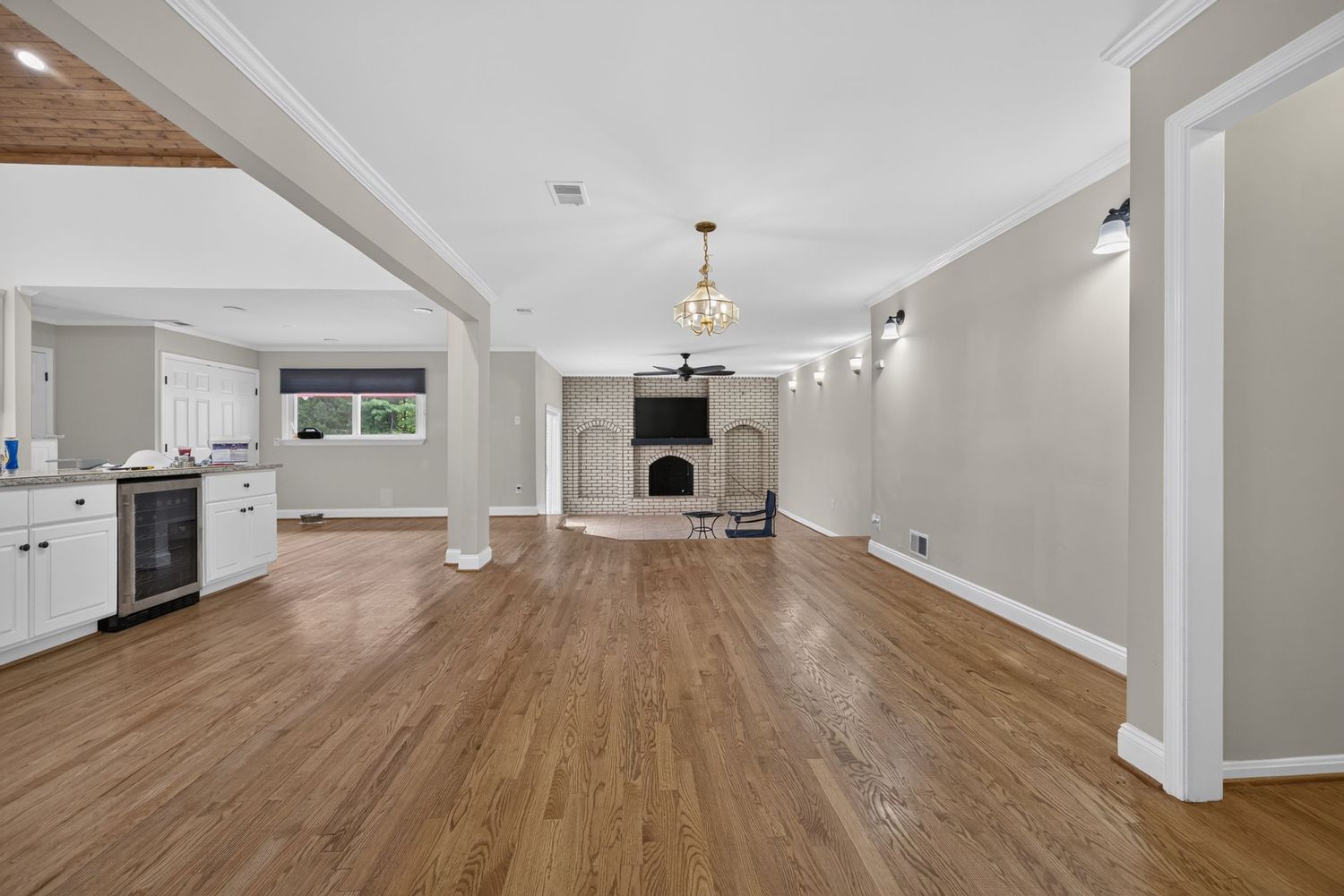 Spacious open-concept living room with hardwood floors, fireplace, chandelier, and white kitchen cabinetry in background.