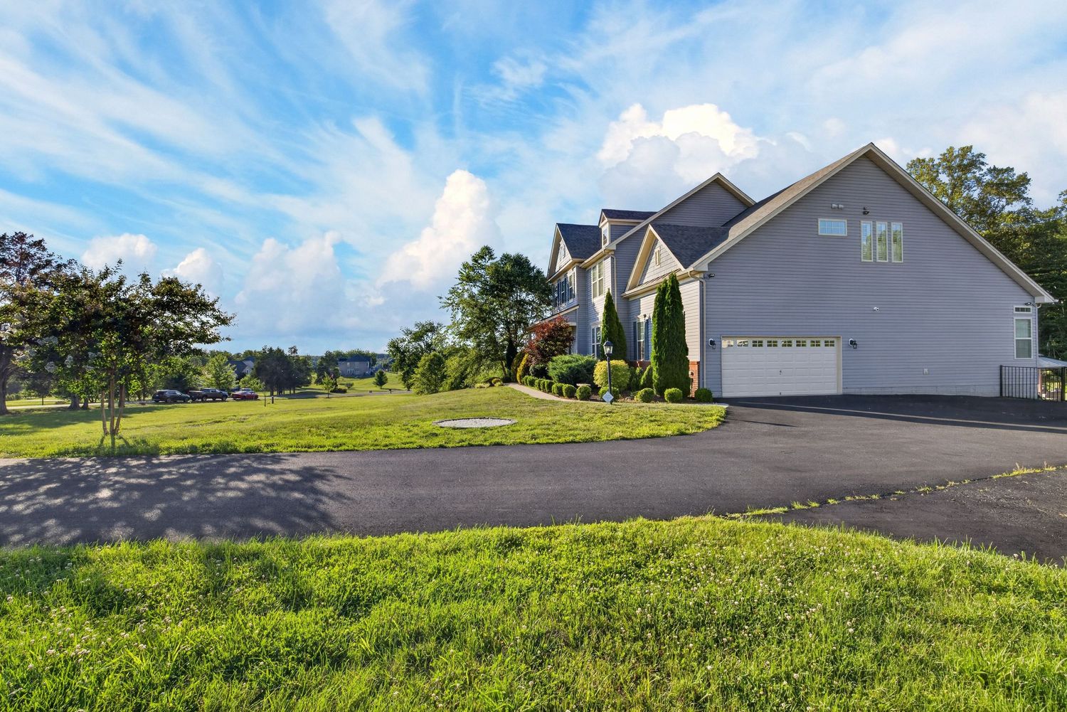 Gray suburban home with attached garage, lush green lawn, paved driveway, and trees under a blue sky with clouds.