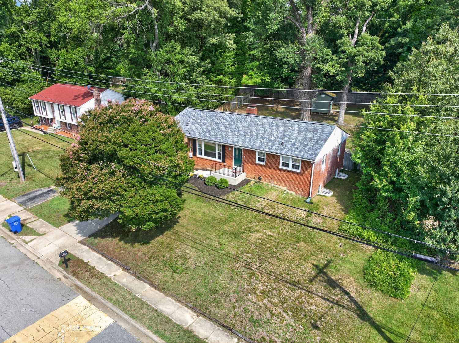 Aerial view of a brick ranch home with gray roof, lush green lawn, and mature trees in a suburban neighborhood.