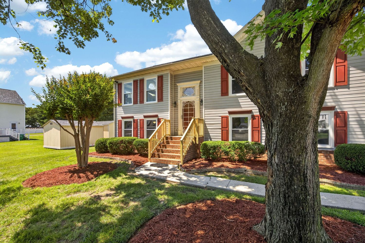 Two-story townhome with red shutters, gold staircase, lush green lawn, and mature trees on a sunny day.