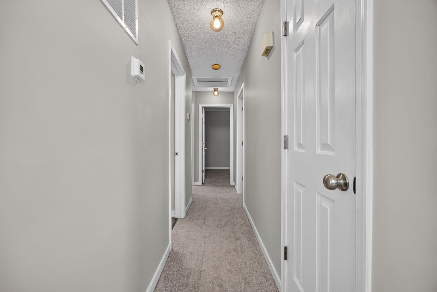 Bright narrow hallway with light gray walls, beige carpet, white doors, and gold ceiling light fixtures.