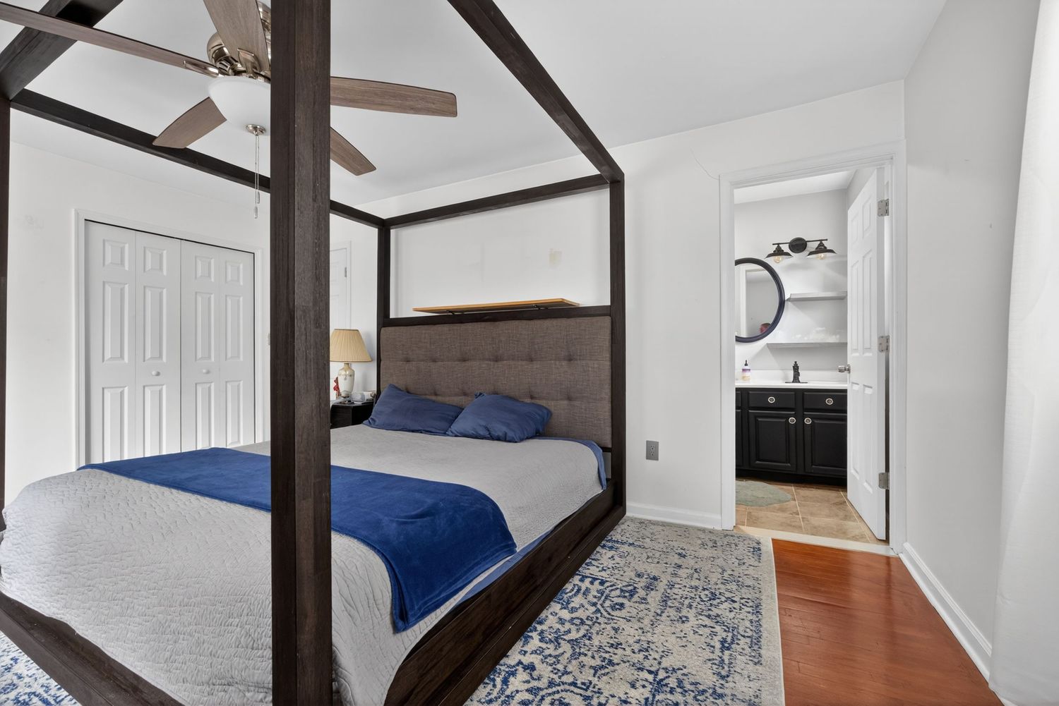 Elegant four-poster bed with blue accents on hardwood floors, featuring an en-suite bathroom and ceiling fan.