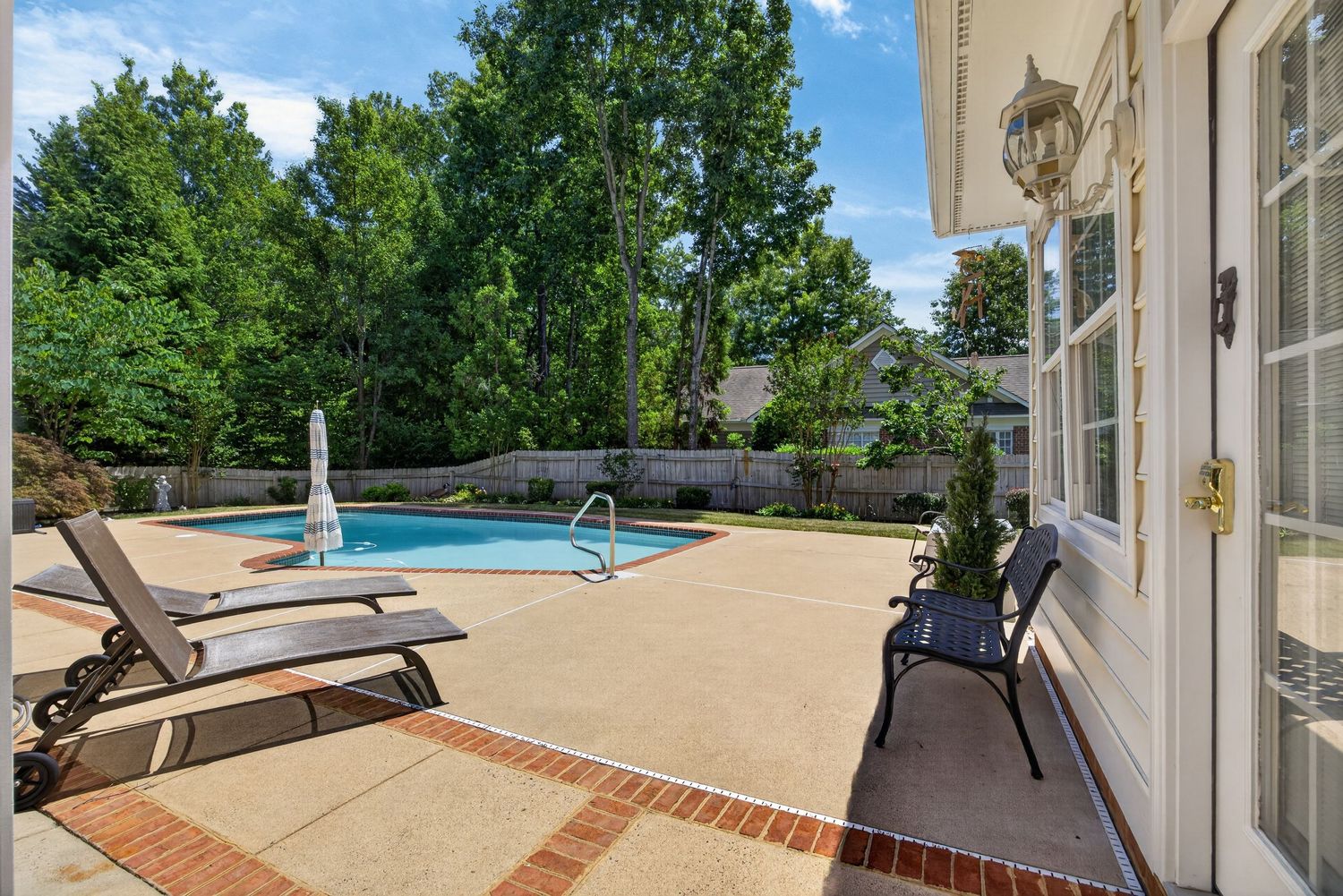 Spacious backyard patio with lounge chairs surrounding a sparkling swimming pool, enclosed by a white fence and lush trees.