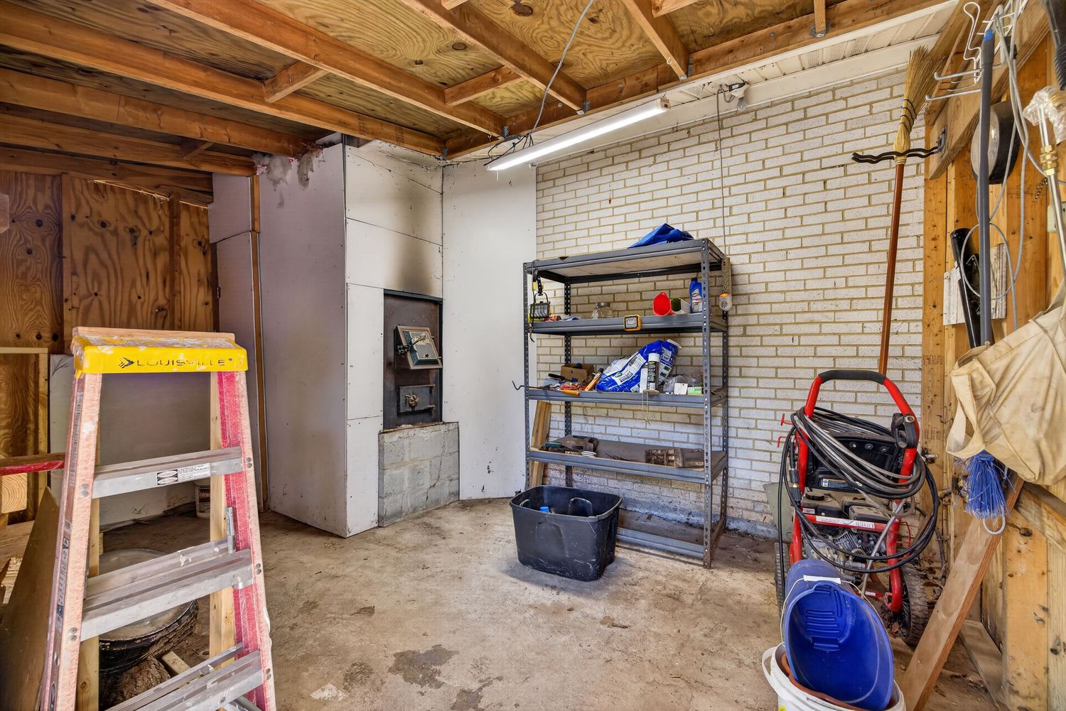 Unfinished garage interior with brick walls, metal shelving unit, yellow ladder, and various tools and equipment on concrete floor.