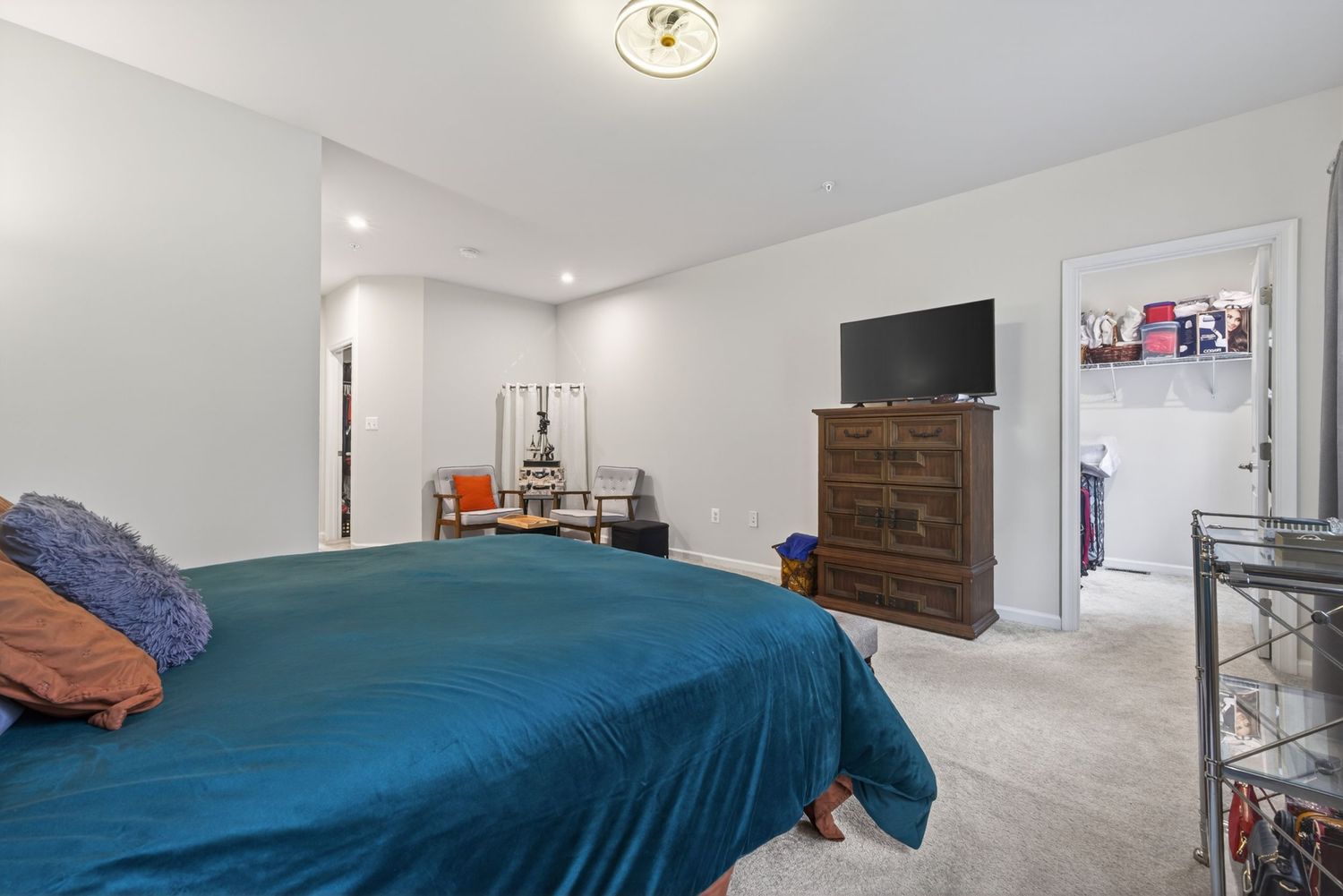 Spacious bedroom with teal bedspread, dresser, wall-mounted TV, and neutral carpet leading to a bright walk-in closet.