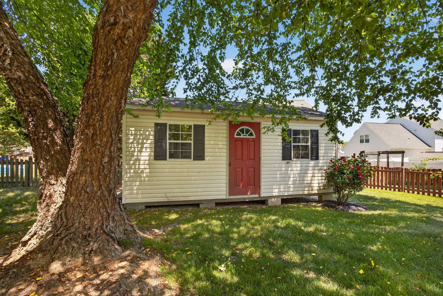 Small yellow cottage with red front door, black shutters, and large shade tree in lush green yard with wood fence.
