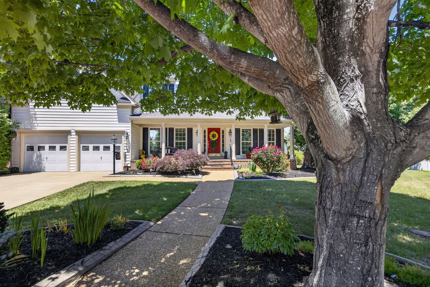Charming suburban home with red front door, covered porch, attached garage, lush lawn, and mature trees lining a concrete walkway.
